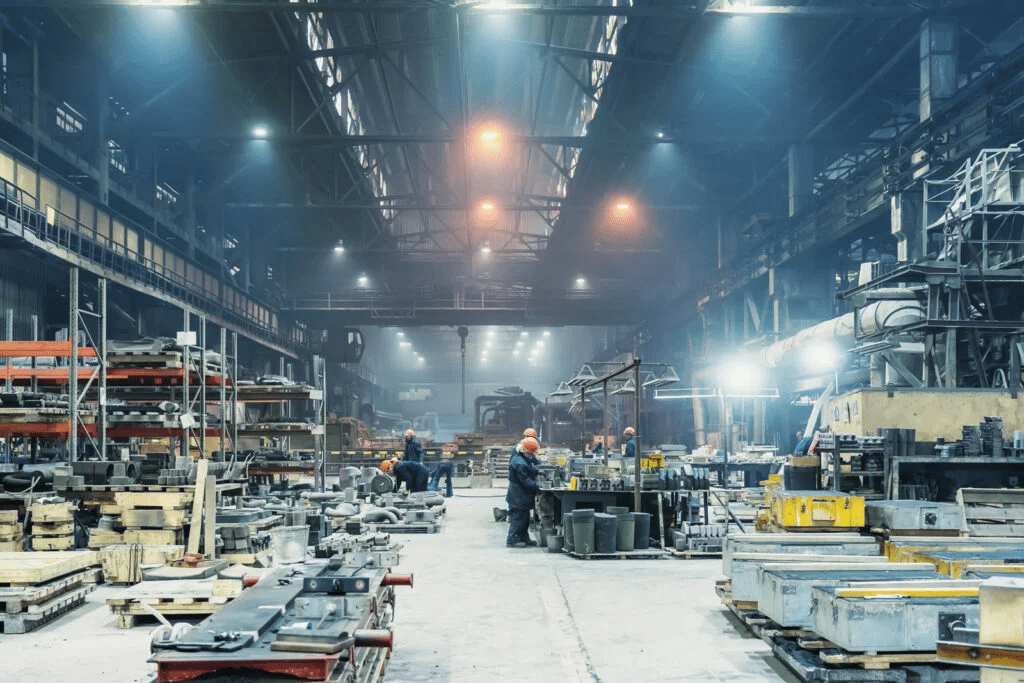 A wide view of a large, modern industrial factory floor, with workers in hard hats and safety gear operating in a bustling manufacturing environment