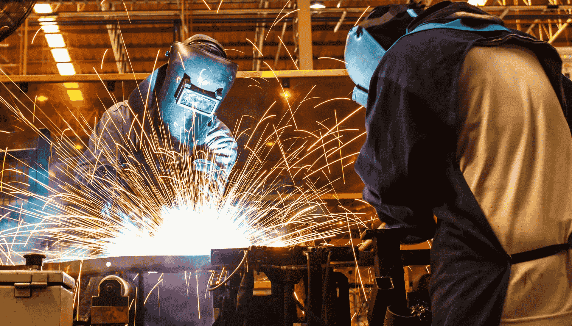 Two welders wearing protective helmets and gear working on a metal project, creating a large shower of bright sparks